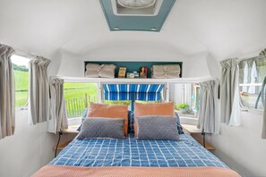 A cozy bedroom with a bed, pillows, a shelf with books and a clock, and a view of a green field. - Airstream, Devon Hideaways - a shepherds hut that sleeps 2 guests in 1 bedroom (Kenn)