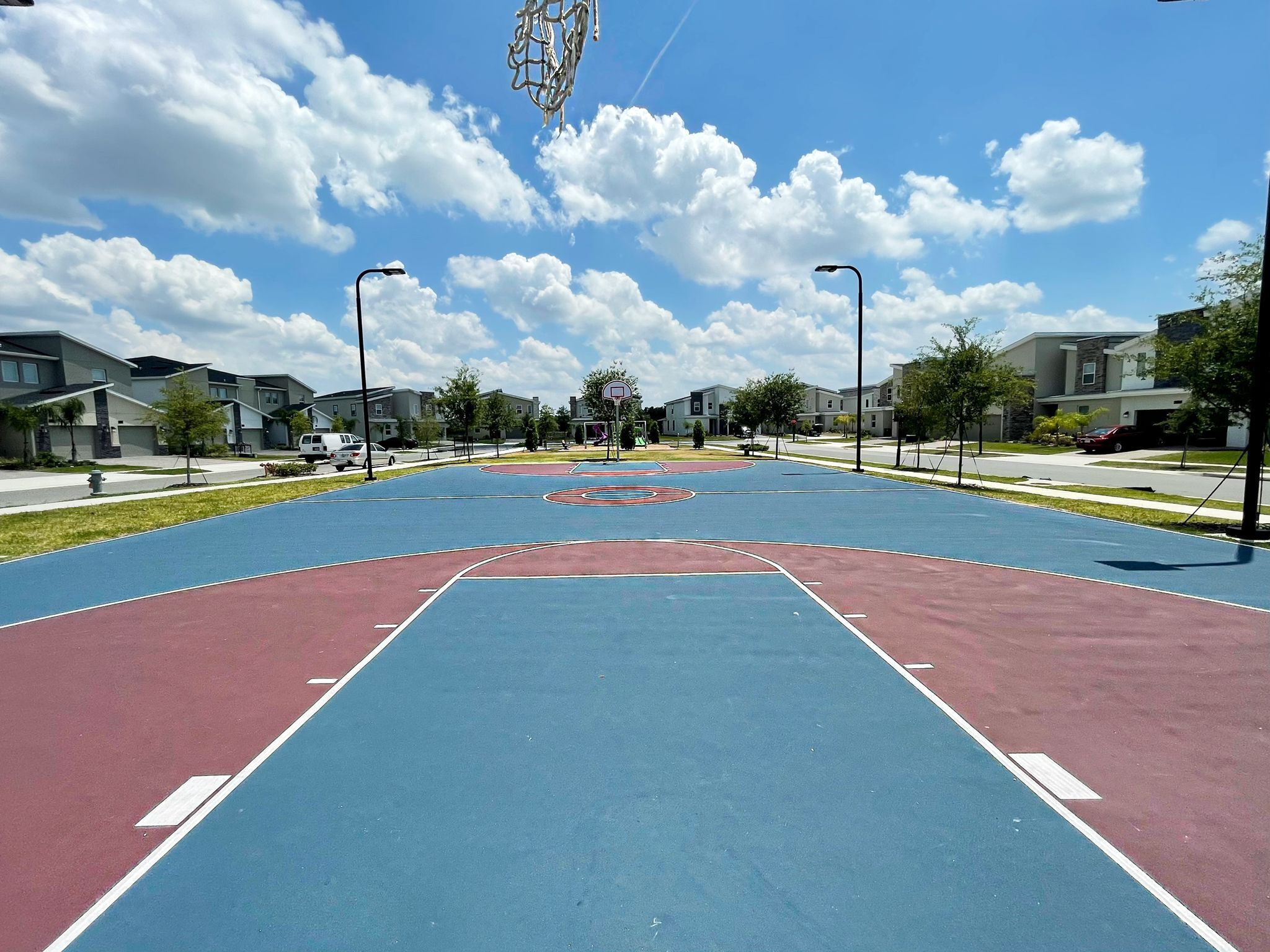 Een basketbalveld buiten met een blauw en rood oppervlak, omgeven door woongebouwen en bomen.