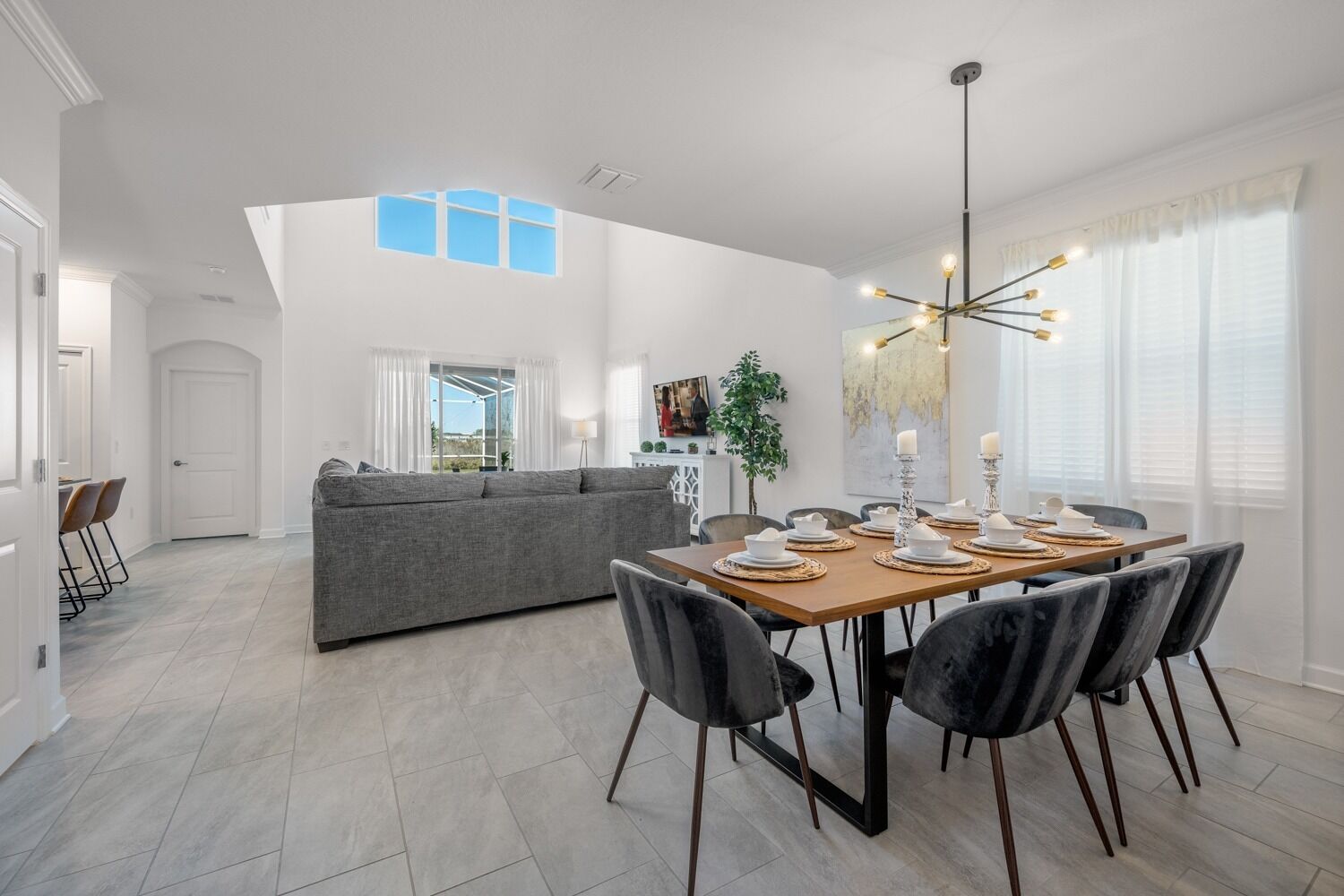 Un comedor moderno con una mesa de madera dispuesta para cuatro personas, rodeada de sillas oscuras. Un sofá gris se encuentra en el fondo, y un ventanal grande proporciona luz natural.