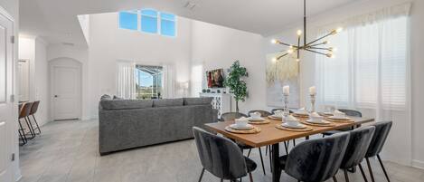 Un comedor moderno con una mesa de madera dispuesta para cuatro personas, rodeada de sillas oscuras. Un sofá gris se encuentra en el fondo, y un ventanal grande proporciona luz natural.