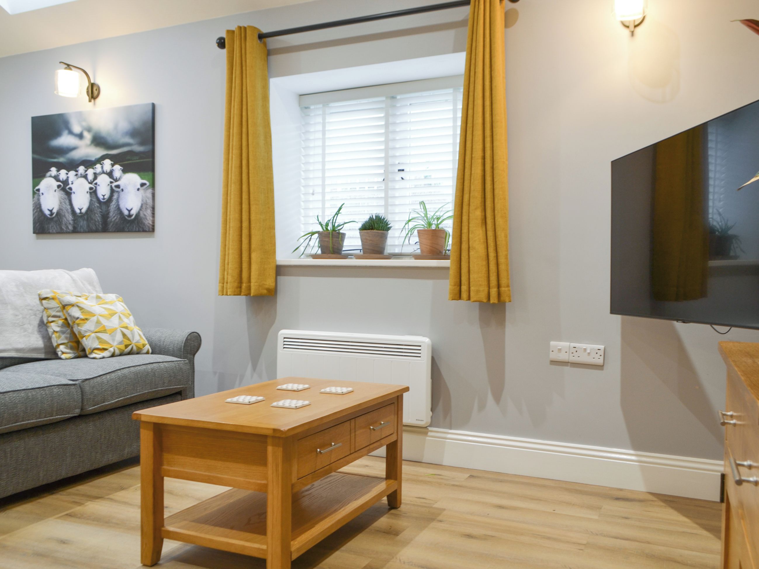 A living room with a sofa, a wooden table, a radiator, a window with curtains, a wall-mounted TV, and a painting.