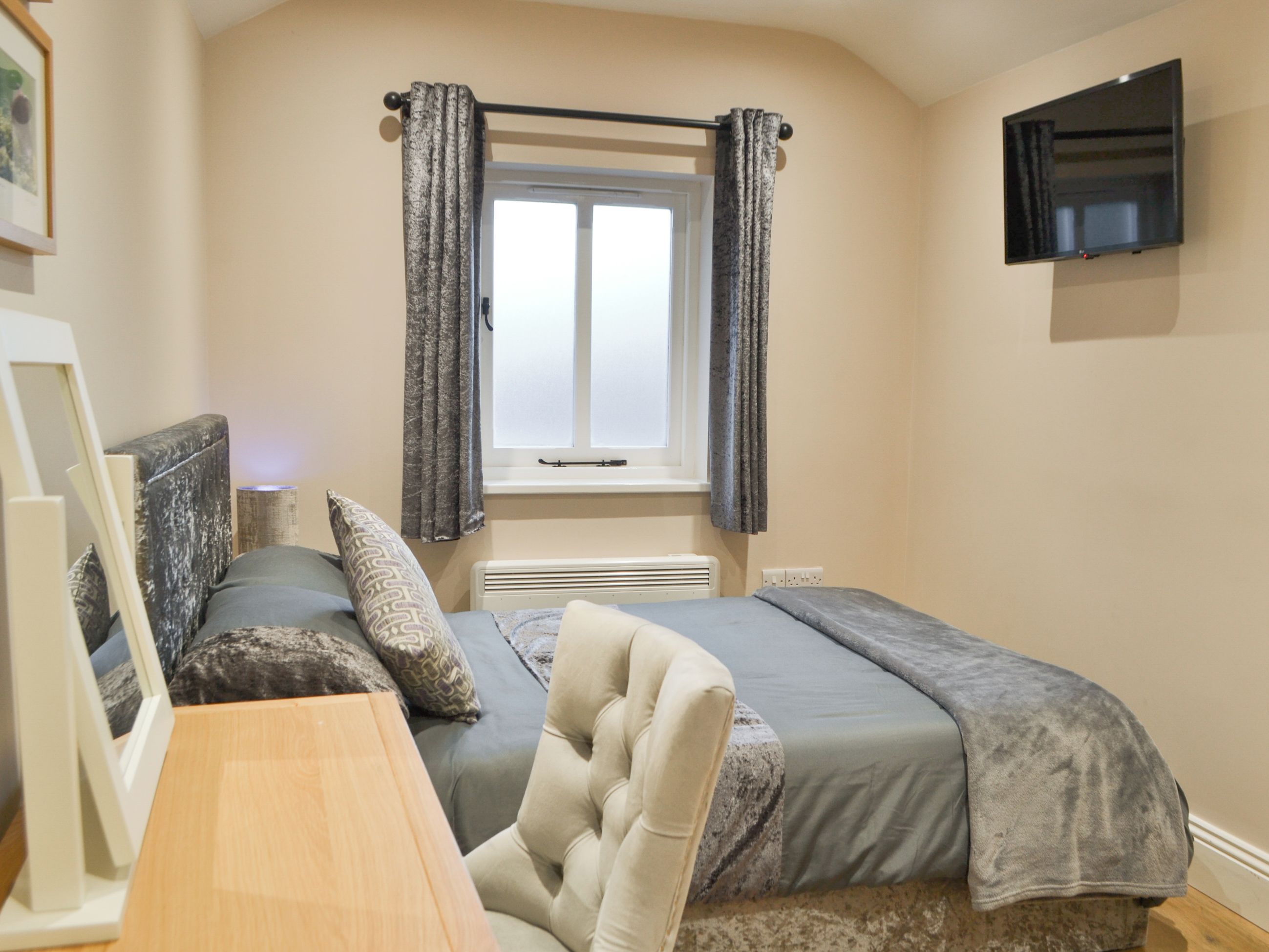 A bedroom with a bed, a desk, a chair, a window with curtains, a flat-screen TV mounted on the wall, and a radiator.