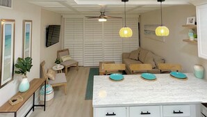A modern kitchen with white cabinetry, a marble countertop, and a central island with teal plates. There are two ceiling fans, a dining area with a wooden table and chairs, and a seating area with a sofa and armchairs. - Pet-Friendly Paradise with Special Discounts - A Wave from It All Awaits!" (Fort Pierce)