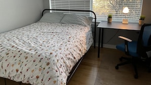Een slaapkamer met een bed, een bureau, een stoel en een raam met jaloezieën.
