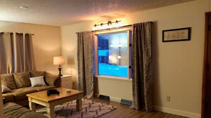 Una sala de estar acogedora con un sofá, una mesa de centro y una lámpara. Se ve una ventana con cortinas, y una cita enmarcada en la pared.