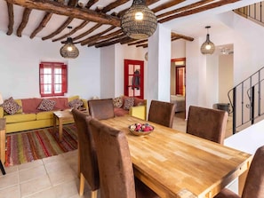 Ein gemütliches Wohnzimmer mit einem Holzesstisch, braunen Stühlen, einem farbenfrohen Sofa und einer roten Tür im Hintergrund.