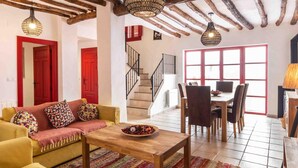 A cozy living room with a sofa, a coffee table, and a dining area with chairs and a table. There is a staircase leading to another level, and a window with red shutters. - Fantastic rural house in Nigüelas (Nigüelas)