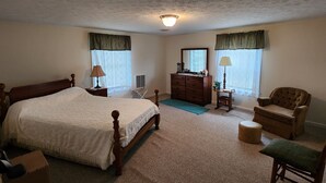 A bedroom with a large bed, dresser, chair, and a small table with a lamp.
