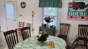 A dining room with a round table, six wooden chairs, a chandelier, a green curtain, a vintage radio, and a sign reading "Natural Farming The Apple House Kitchen".