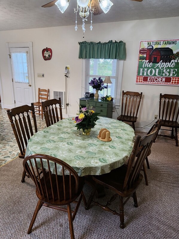 Comedor con una mesa redonda, seis sillas de madera, una araña, una cortina verde, una radio antigua y un letrero que dice "Natural Farming The Apple House Kitchen".