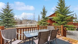 Une terrasse en bois avec une table et des chaises en verre, entourée d’arbres et avec un chalet en arrière-plan.