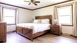Un dormitorio con cama de madera, mesita de noche, ventilador de techo y ventanas grandes con cortinas.