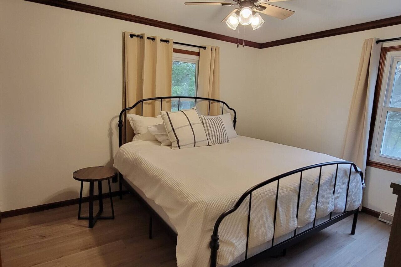 A bedroom with a bed, a small stool, a ceiling fan, and a window with curtains.