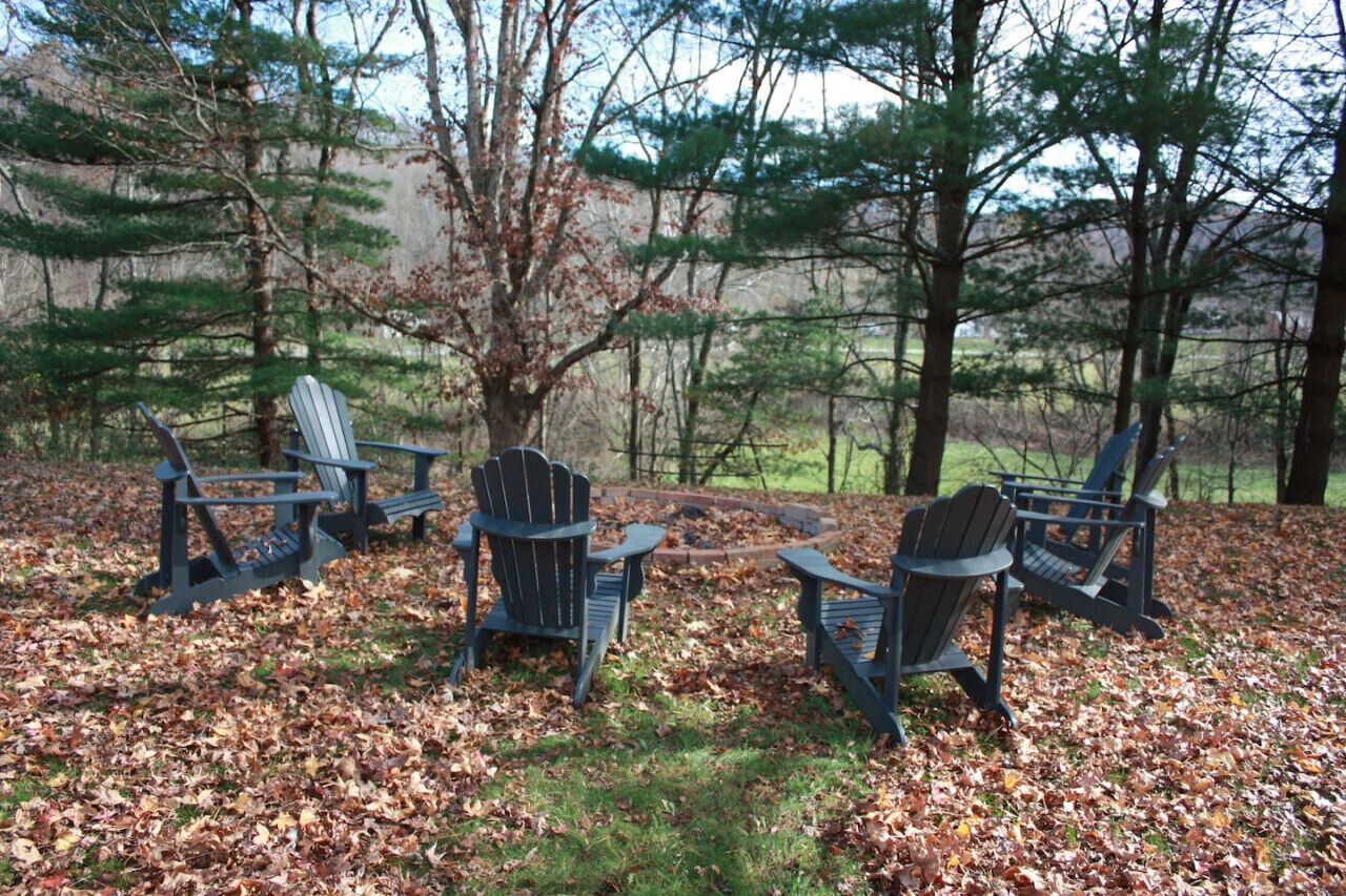 Four Adirondack chairs arranged in a circle on a leaf-covered ground with trees in the background.
