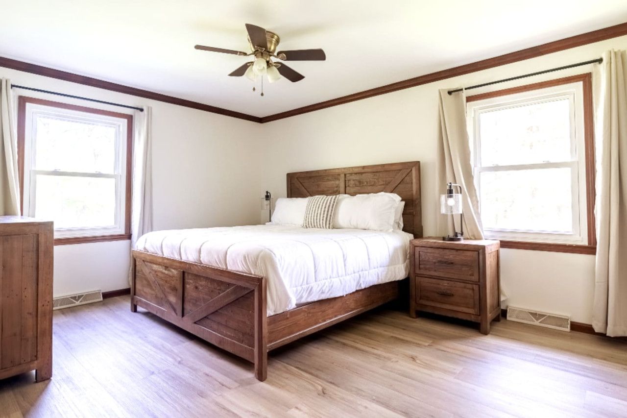 A bedroom with a wooden bed, a nightstand, a ceiling fan, and large windows with curtains.