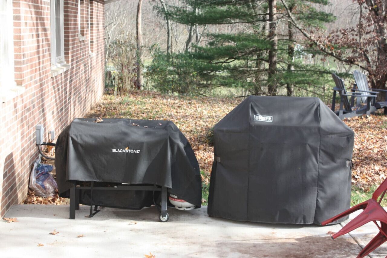 Deux barbecues d’extérieur, recouverts de housses de la marque BLACK+DECKER, sont placés sur une terrasse en béton, avec un bâtiment en briques et des arbres en arrière-plan.