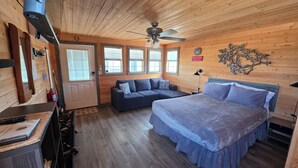 Una habitación de cabaña de madera con cama, sofá, ventilador de techo y ventana con persianas.