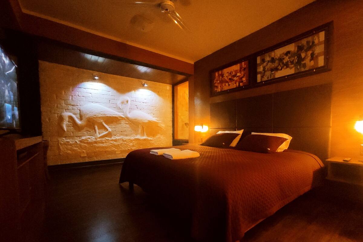 Una habitación con poca luz, una cama, una pared con una escultura de luz y un ventilador de techo.