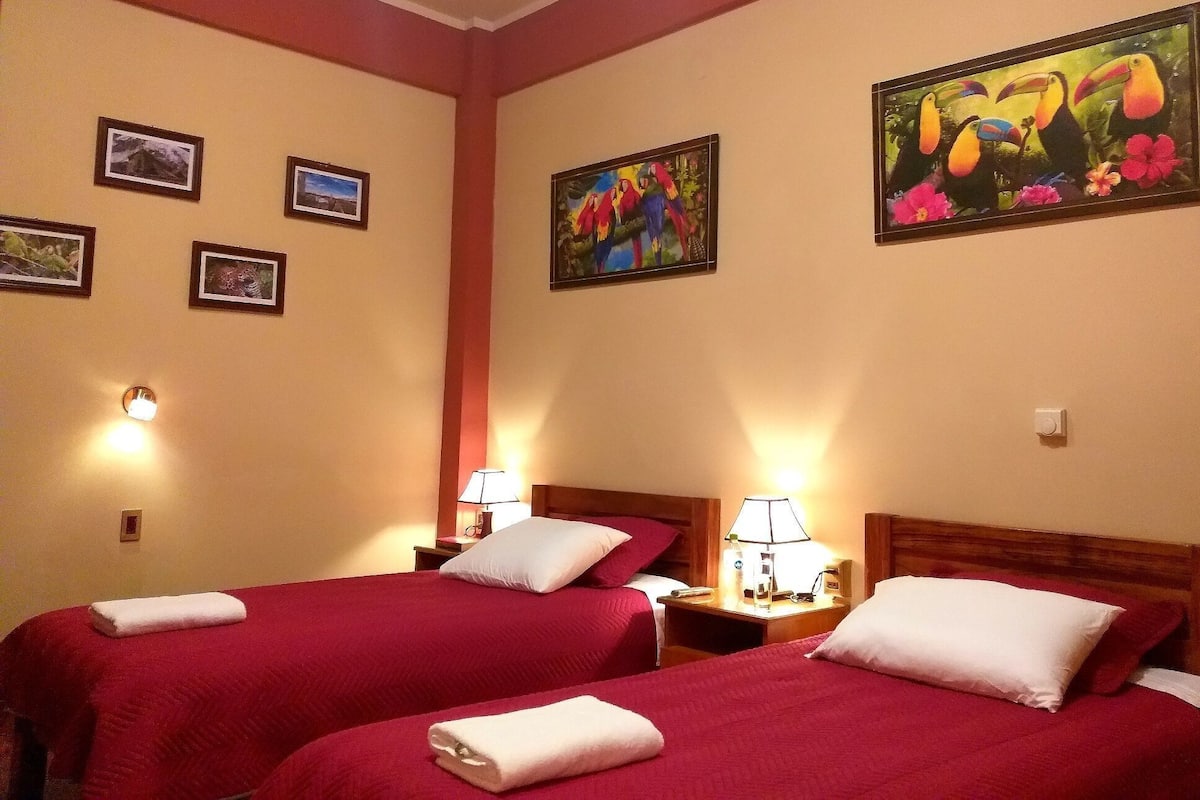 Habitación de hotel con dos camas, cada una con cubrecama rojo y almohadas blancas, una mesita de noche con lámpara y una obra de arte en la pared.