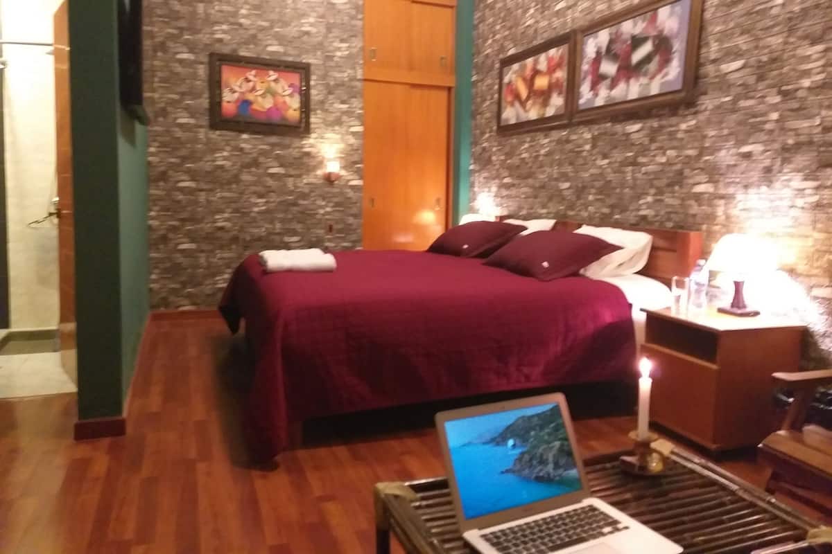 Un dormitorio con cama, una laptop sobre una mesita auxiliar, una mesita de noche de madera con lámpara y pared con textura de piedra.