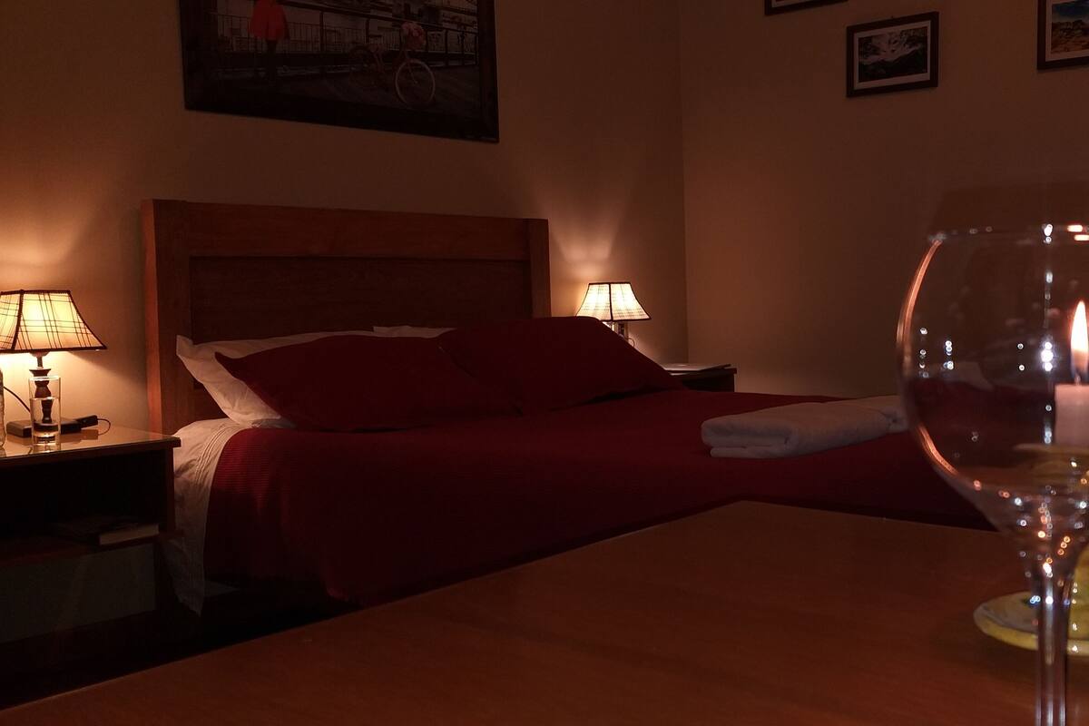 Una habitación de hotel con una cama, una mesita de noche con una lámpara, una botella de vino y una copa de vino sobre una mesa.
