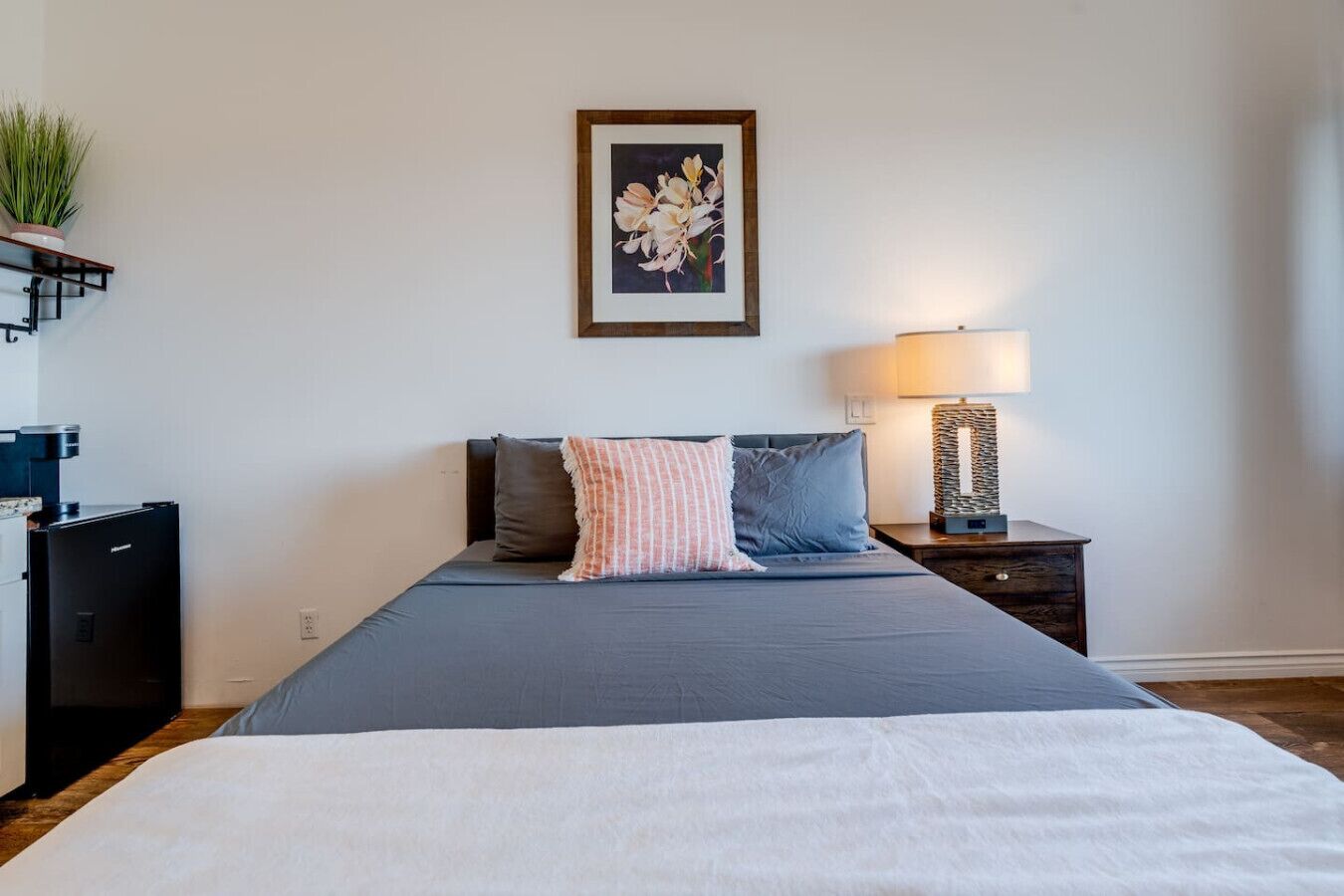 A bedroom with a bed, pillows, a bedside table with a lamp, a small plant on a shelf, and a coffee machine.