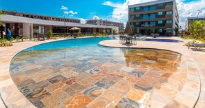 A swimming pool with a unique tile design, surrounded by outdoor seating and a modern building in the background. - Mana Beach Experience B221 por Carpediem (Ipojuca)