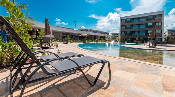 A pool area with lounge chairs and a building in the background. - Mana Beach Experience B221 por Carpediem (Ipojuca)