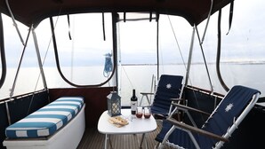A boat deck with a cushioned bench, a small table with a bottle and glasses, and two lounge chairs. - Paradise on the Great South Bay (Cherry Grove)