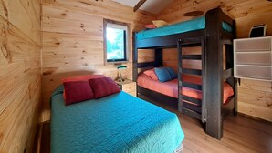 Une chambre dans un chalet en bois, avec un lit superposé, un lit simple recouvert d’une couverture rouge et une fenêtre donnant sur la forêt.
