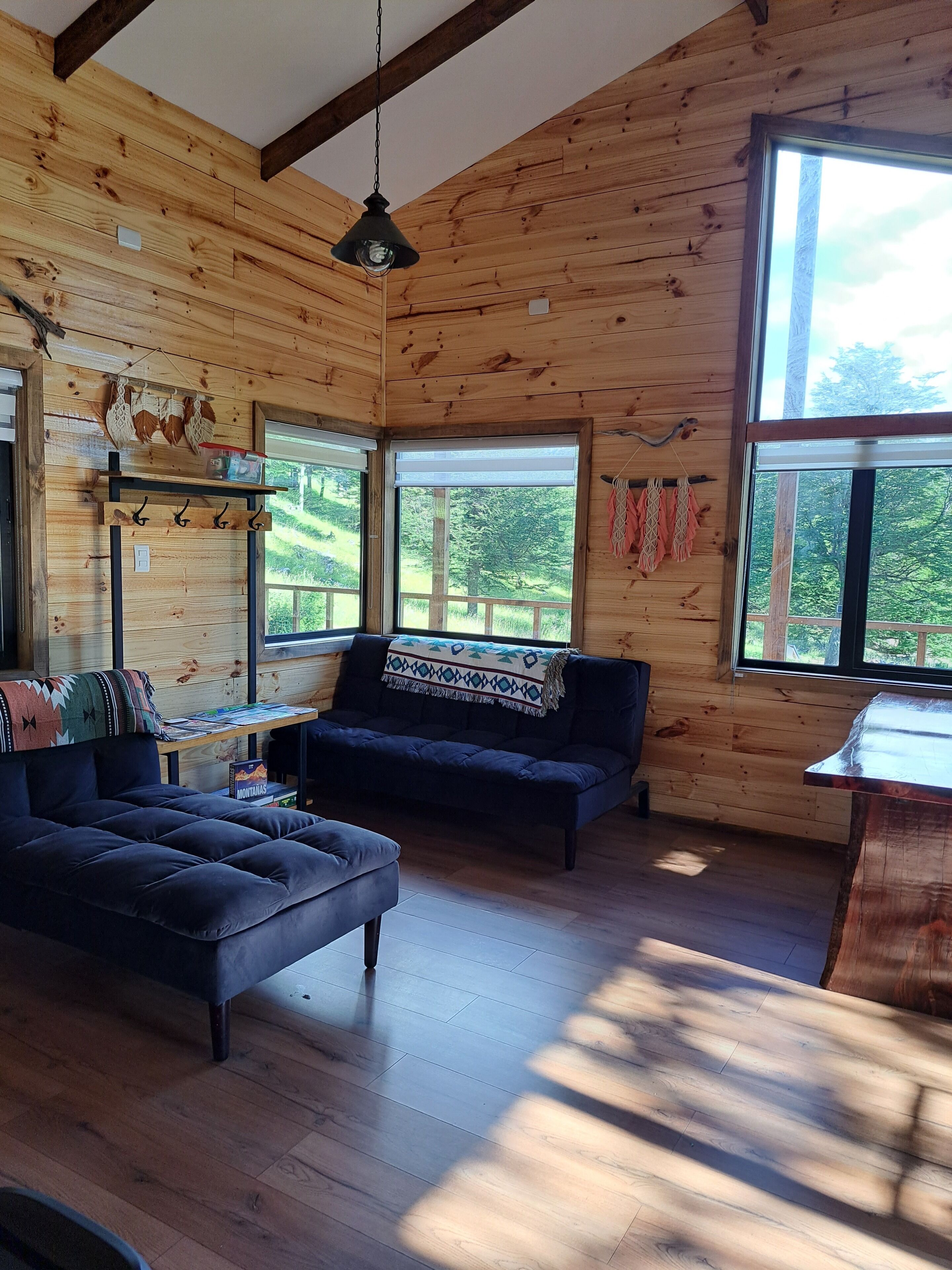 A cozy cabin interior with wooden walls, a dark sofa, a table with a blanket, and large windows offering a view of the outdoors.