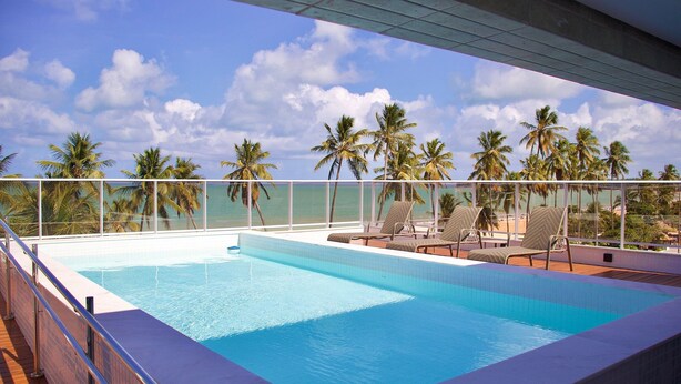 A swimming pool with lounge chairs and a view of palm trees and the ocean. - Apart na Orla de Cabo Branco - Carpediem (João Pessoa)