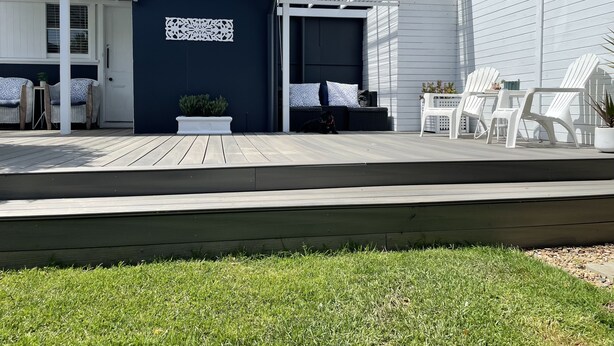 Un patio exterior moderno con cubierta de madera, sillas blancas, una mesita y una planta en maceta.