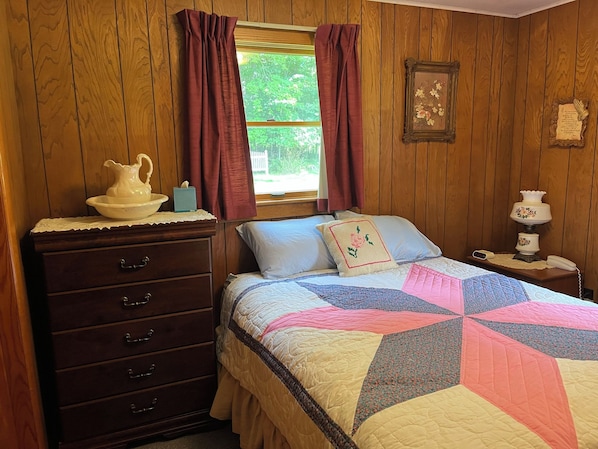 A bedroom with a wooden paneled wall, a bed with a quilt, a nightstand with a lamp, a dresser, and a window with curtains.