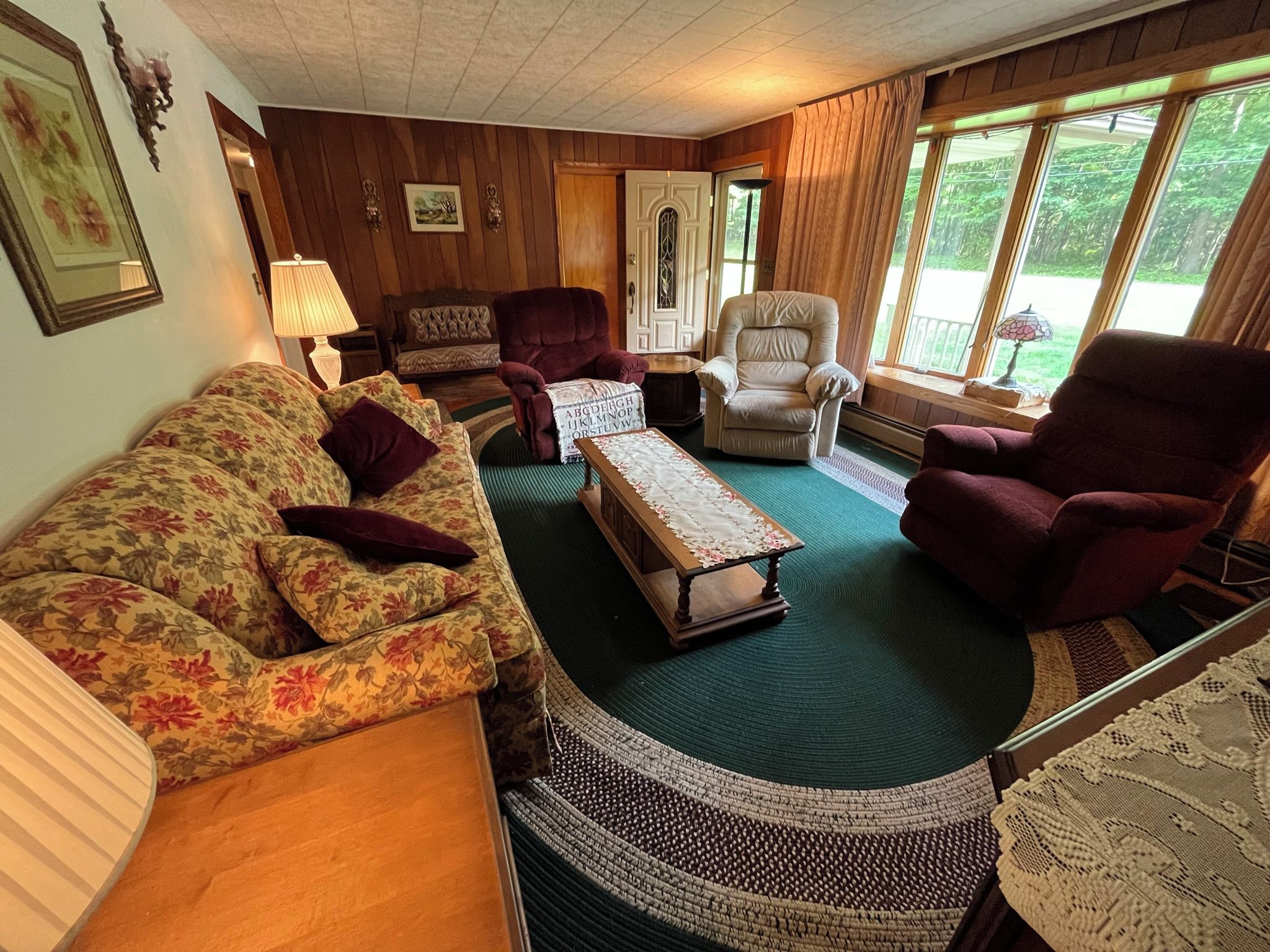 Una sala de estar acogedora con un sofá floral, dos sillones, una mesa de centro y vistas a la vegetación a través de amplias ventanas.
