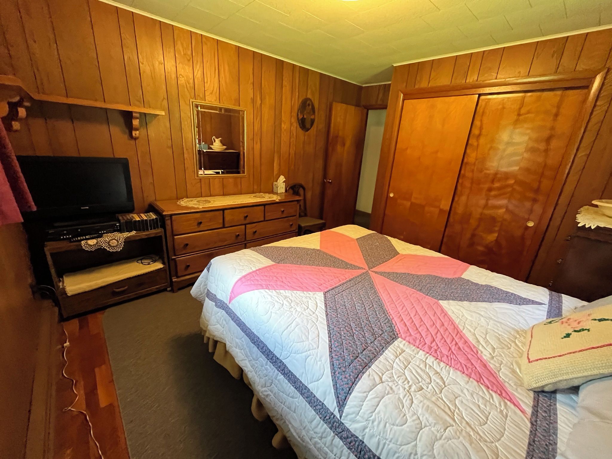 Un dormitorio con paredes de madera, televisor, cómoda, cama con colcha y armario.