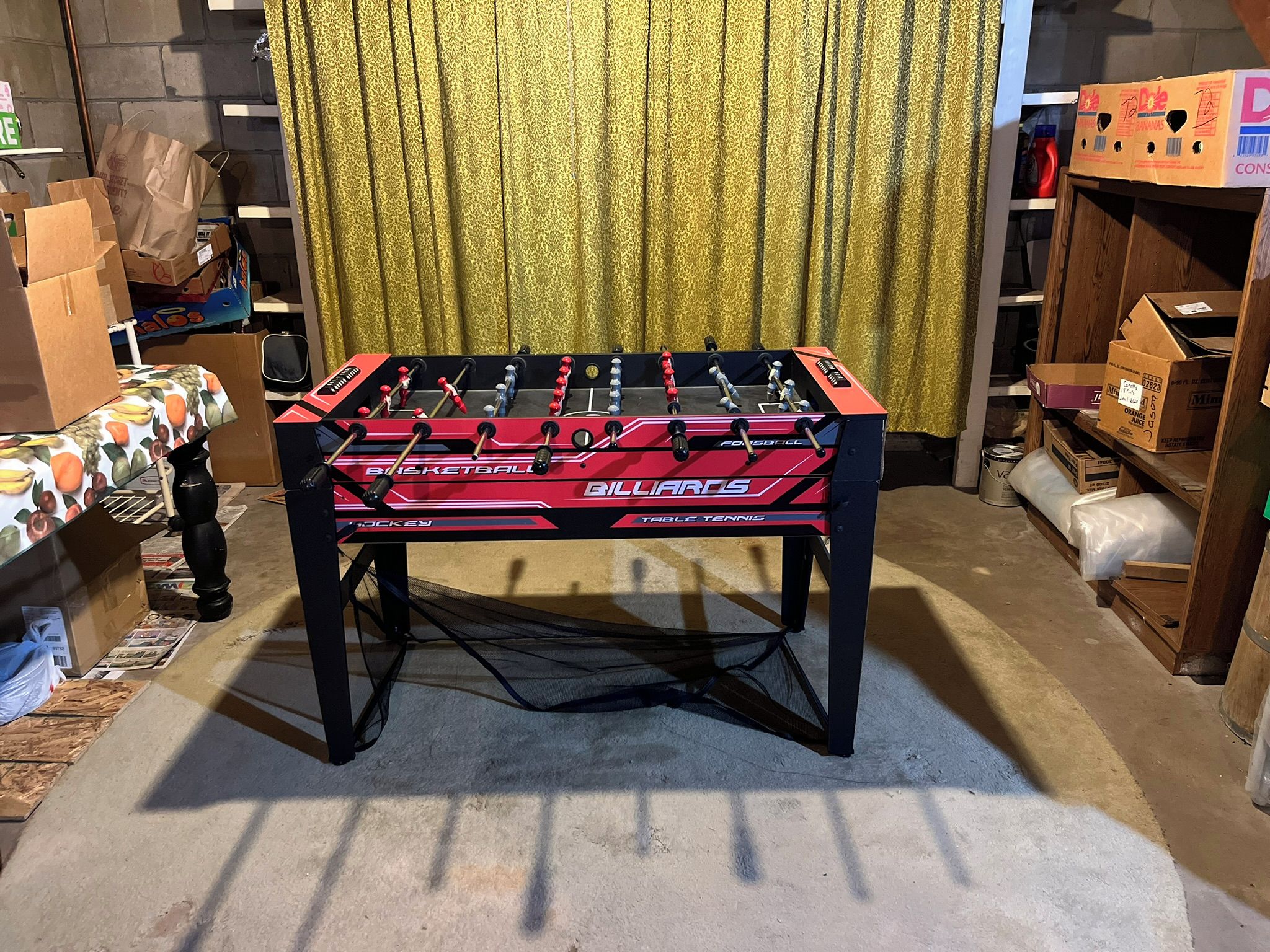 Une table de baby-foot dans une pièce où l’on aperçoit un rideau à motifs et divers cartons et objets en arrière-plan.