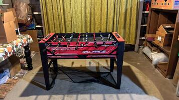 Une table de baby-foot dans une pièce où l’on aperçoit un rideau à motifs et divers cartons et objets en arrière-plan.