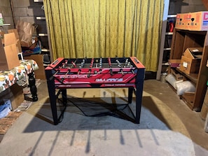 A foosball table in a room with a patterned curtain and various boxes and items in the background.