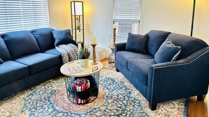 Un salon avec deux canapés bleu foncé, une table basse ronde surmontée d’un tapis à motifs et un grand lampadaire.