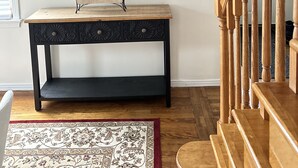 Un banc en bois doté d’une structure métallique et d’une assise tissée, une table en bois foncé avec des tiroirs, et un tapis à motifs posé sur un plancher en bois.