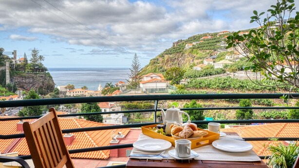 Un balcón con una mesa preparada para el desayuno, con vistas a un pueblo costero de tejados de terracota y vista al mar.