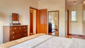 Un dormitorio con un aparador de madera, una cama con colcha estampada, un espejo y una puerta que da a otra habitación.