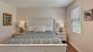 Une chambre à coucher comprenant un lit, deux tables de chevet, un ventilateur de plafond, une fenêtre avec des stores et un tableau accroché au mur.
