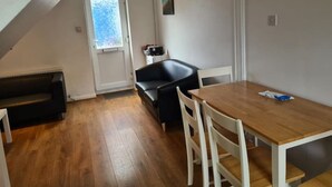 Una habitación pequeña y acogedora con una mesa y sillas de madera, un sofá de cuero negro y una puerta blanca con ventanal.