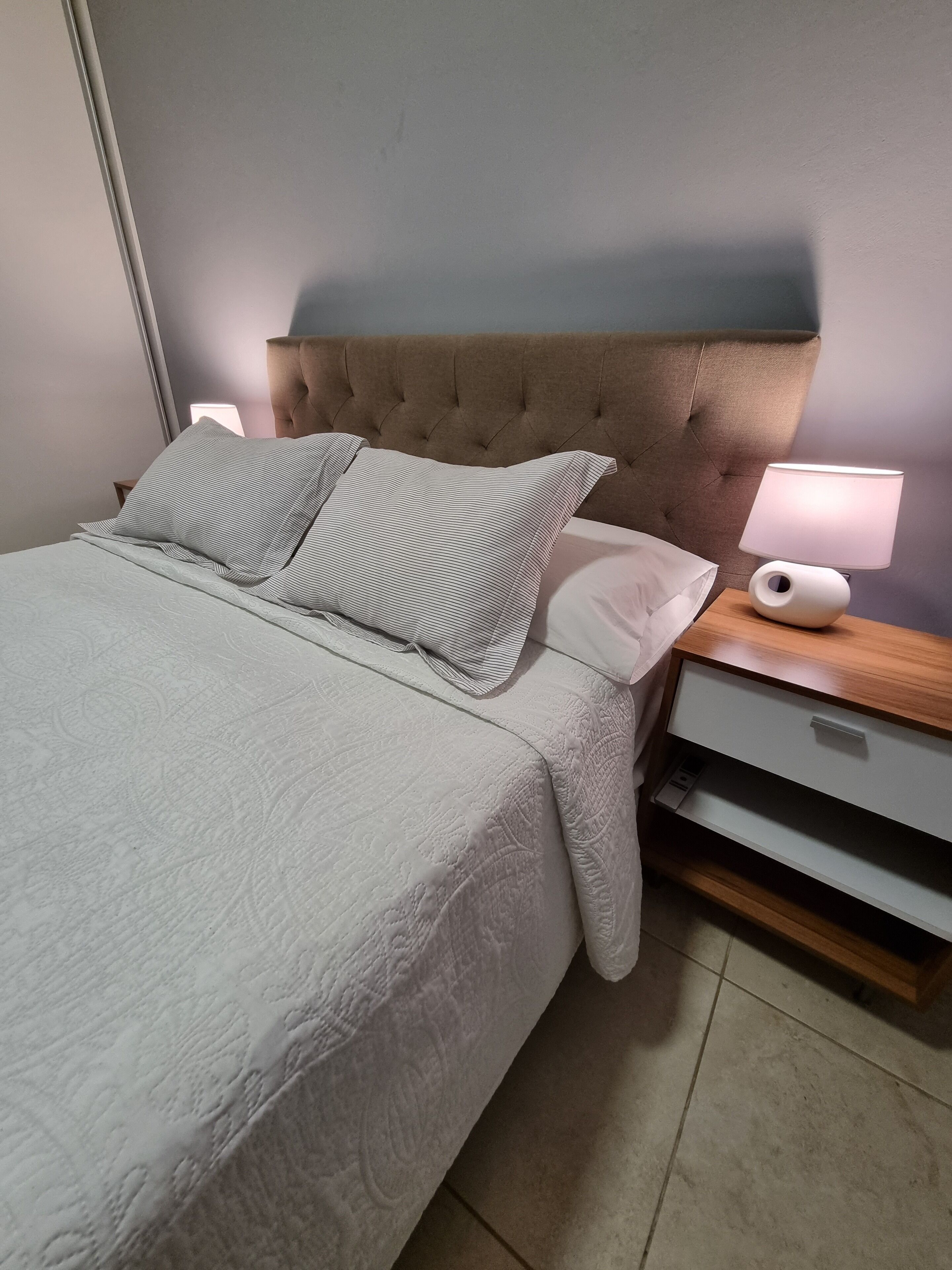 Un lit bien fait, avec une tête de lit capitonnée, deux oreillers, une table de chevet avec une lampe et une petite table de nuit avec un tiroir.