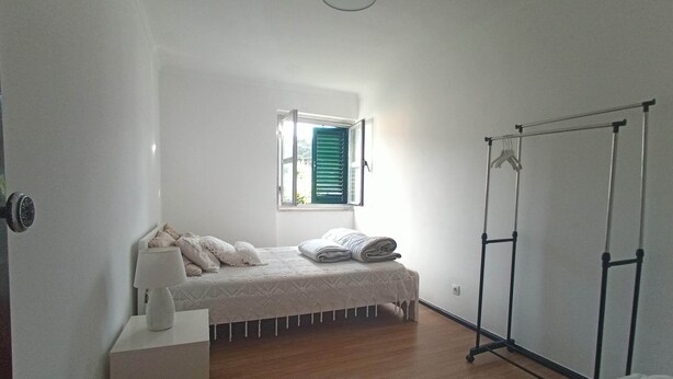 A bedroom with a bed, a nightstand, a window with blinds, a wardrobe, and a ceiling light. - Ginjals-67 (Almargem do Bispo)