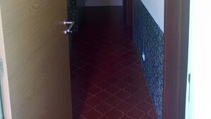 A hallway with a wooden door on the left, a white door in the center, and a wooden door on the right. The floor has a red patterned tile. - Ginjals-67 (Almargem do Bispo)