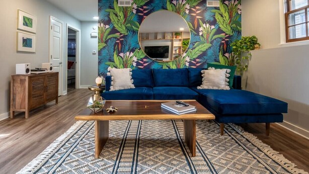 Ein Wohnzimmer mit einem blauen Ecksofa, einem runden Spiegel, einem Couchtisch und einem gemusterten Teppich.