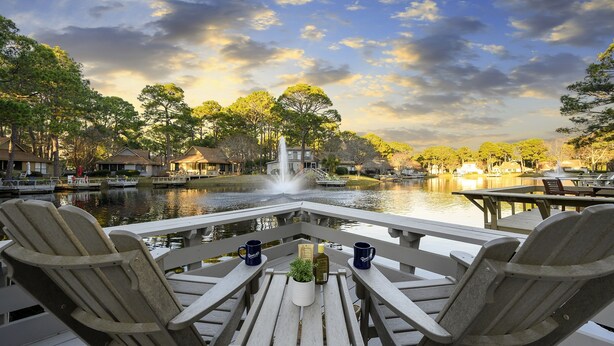 A wooden deck with Adirondack chairs and a table, overlooking a calm river with a fountain and boats. - Lakefront Sandestin Home w/ Golf Cart &Arcade Room (Mir)